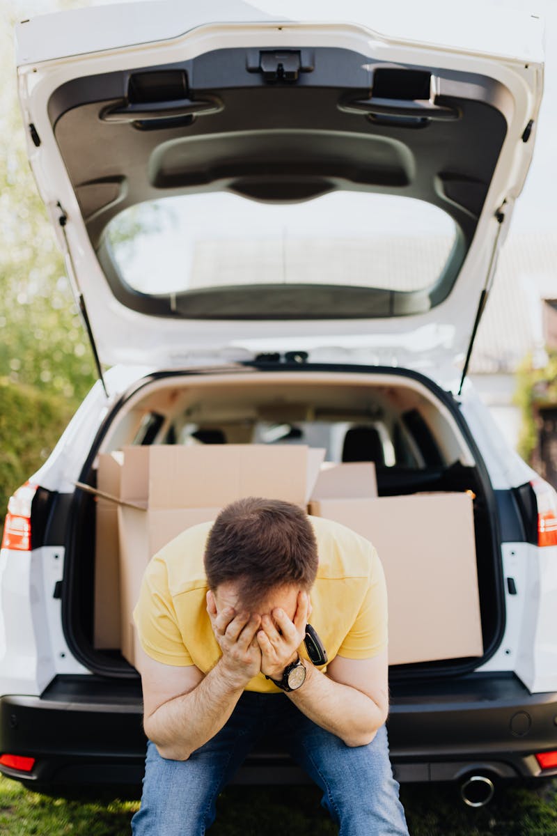 Man sitting by car with head in hands, surrounded by moving boxes, feeling stressed.